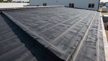 A high-angle professional architectural photograph of a perfectly installed dark charcoal gray EPDM rubber roof on a clean, modern commercial building in New Jersey. The sky is clear, casting sharp daylight that highlights the technical precision of the waterproof seams. North American / US construction setting, showing durability and industrial quality.