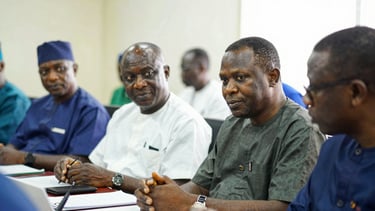 A medium close-up documentary photo of Nigerian lawmakers in discussion inside a brightly lit committee room, natural daylight.