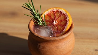 Vibrant drink photography of a mezcal cocktail in a custom clay cup, garnished with a slice of blood orange and a sprig of rosemary, warm golden-hour lighting, rich textures, shallow depth of field