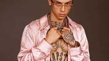 Portrait of Central Cee wearing a pink silk shirt, gold chains, and glasses with visible tattoos.