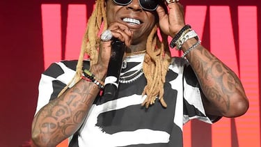 Rapper Lil Wayne performing on stage with a microphone, wearing sunglasses and a black and white shirt.