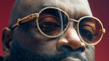Close-up of Rick Ross wearing diamond-encrusted sunglasses and exhale cigar smoke.