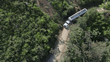 Aerial view of a white fuel tanker truck driving on a narrow dirt road through a dense tropical forest.