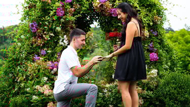 Man proposing on one knee to a woman in a garden setting, holding a ring and a bouquet of red flower