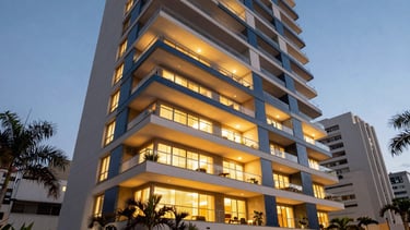 A low-angle professional architectural photograph of a modern residential building in a South American / Brazilian city during the blue hour. The building features sleek balconies with warm gold light emanating from within, clean white and dark blue exterior lines, and premium landscaping with tropical plants in the foreground under soft, sophisticated lighting.