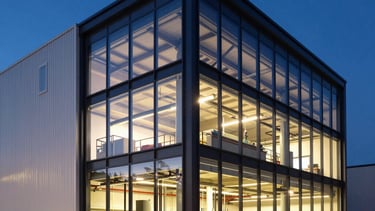 A professional architectural photography shot of a modern European industrial facility facade at dusk. The building features clean glass and dark steel lines, with warm interior light highlighting the CNC machinery area inside. The sky is a deep midnight blue, contrasting with the sharp midnight blue and off-white exterior.