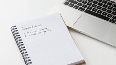 A person wearing a Vancouver Police uniform is partially obscured by greenery. The individual is wearing a black cap and jacket with 'Vancouver Police' insignia. Raindrops are visible on the clothing, indicating wet weather conditions. The background is blurred, featuring a building and possibly another person at a distance.