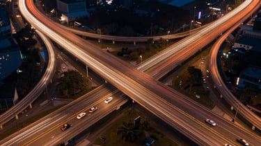 Drone photography of a complex highway interchange at night, with light trails from cars creating vibrant orange and white lines against a dark blue background.