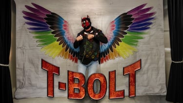 A man poses in front of a mural wearing a pup mask. Name plate: T-BOLT