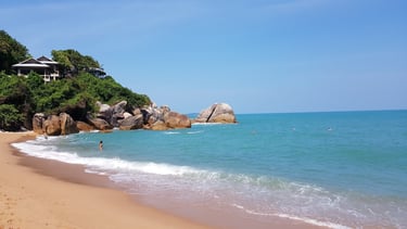 Tropical beach in Koh Samui with turquoise water, granite boulders, and a luxury villa on a lush hillside.