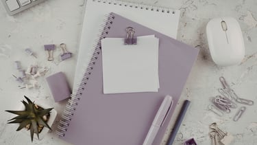 Flat lay of purple office supplies including spiral notebooks, a white mouse, and stationery on a desk.