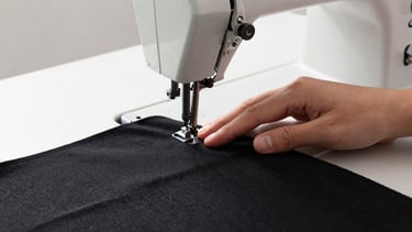 Hands of a tailor carefully guiding heavy black cotton fabric through a vintage sewing machine, soft side light, industrial workspace