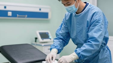 A professional cleaning technician in a modern medical setting in North American / US, wearing a face mask and protective blue gear, meticulously sanitizing a hospital exam table with a cloth. The background is a sterile, bright clinical room with light green and dark blue accents. Clean and sharp photography.