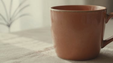 Close-up macro shot of a warm ceramic mug on a textured linen surface, soft morning light creating gentle shadows, abstract botanical line art visible in the background, muted terracotta and cream tones, shallow depth of field, 35mm film texture.