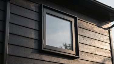 Close-up of a modern matte black window frame installed in a dark siding wall, crisp morning sun casting sharp shadows, 35mm