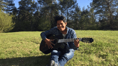 Roger Bardales playing an acoustic guitar while sitting on grass in a sunny park in Lithuania.