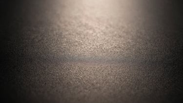 Toma macro de la superficie de una máquina de afeitar mate-negra, mostrando la textura finamente granulada del material. Iluminación lateral dramática, alto contraste, reflejos cálidos y sutiles.