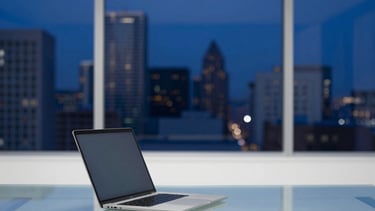 A high-end, minimalist creative agency office in a North American / European / Middle Eastern setting. The scene features a sleek glass desk with a slim laptop, overlooking a blurred city skyline at dusk. The lighting is sophisticated, emphasizing deep navy and royal blue tones with crisp white highlights and a clean, professional aesthetic.