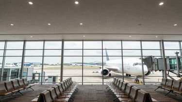 Wide overhead shot of an airport boarding gate interior, a commercial aircraft visible through the floor-to-ceiling glass in the right third of the frame, neutral terminal lighting, clean lines of seating rows in the foreground, expansive negative space of pale terminal ceiling above
