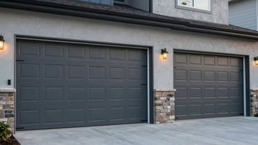 A professional wide-angle photograph of a modern North American residential home with two charcoal gray, insulated garage doors and high-quality hardware. The scene is shot in soft afternoon light on a clean concrete driveway, emphasizing reliability and professional craft. The color palette features slate blue and light gray tones.