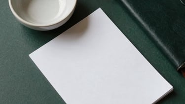 Top-down view of an organized desk with a ceramic bowl, a glass paperweight, and a leather-bound journal. Paper white and charcoal green colors. Minimalist and serene South Asian professional workspace.