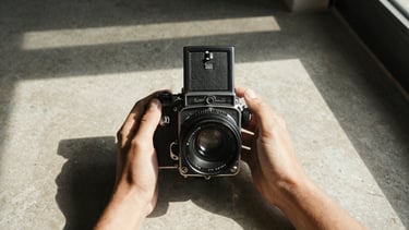 Hands holding a vintage medium-format camera, harsh direct sunlight casting deep geometric shadows on a warm concrete surface.