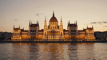 Cinematic wide shot of the Hungarian Parliament building from across the Danube river at sunset, glowing golden lights reflecting on the water, editorial 35mm look