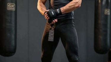 A cinematic, high-performance photograph of a male combat athlete wrapping his hands in a dark, premium MMA gym. He is wearing long black training leggings that fully cover the knees and a dark grey compression top. Global / Performance Athletics. The lighting is dramatic and moody, with gold and off-white highlights on his silhouette. The background is a clean, masculine space with heavy bags and dark grey walls.