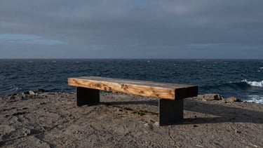 Wide panoramic view of a sculptural bench made of heavy timber and steel, placed on a rugged coastal cliff overlooking a stormy sea, moody overcast twilight, low-angle heroic shot, 35mm lens