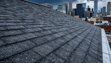 A low-angle professional photograph of a reinforced high-durability roof with dark gray storm-resistant shingles. Rain droplets are visible on the surface under a dramatic navy blue storm sky over the New York City skyline. The lighting is moody, highlighting the structural strength of the materials. North American / US - New York City.