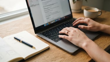 Close-up of hands typing on a laptop on a clean wooden desk under soft natural window light, an open notebook with gold pen nearby, 35mm editorial photography