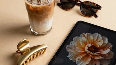 An aesthetic flat lay of a chic morning setup on a warm beige table. A glass of iced latte, a gold hair claw clip, a pair of stylish tortoise-shell sunglasses, and a digital tablet showing a floral screensaver. North American / US lifestyle.