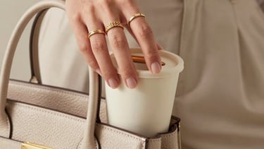 A close-up shot of a chic mom's hand wearing elegant gold rings, lifting a cute neutral-colored toddler snack cup from a premium leather bag. Bright, natural, high-fashion mom style in North American / US.