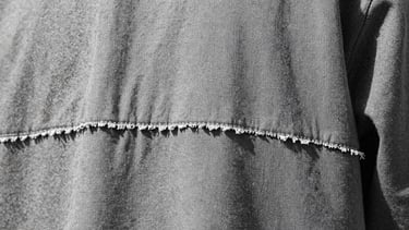 Extreme macro crop of raw-edged hem stitchwork on a heavy-gauge canvas garment, cold overcast daylight, sharp focus, brutalist aesthetic.