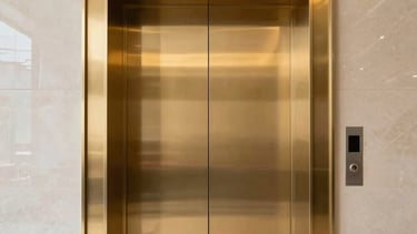 A low-angle, professional architectural photograph of a sleek, modern elevator entrance in a South Asian corporate headquarters. The doors are finished in polished gold, reflecting a minimalist lobby with black marble floors and soft, premium lighting. The composition is centered and symmetrical, projecting corporate reliability and precision.