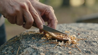 Extreme close-up of hands carving a wooden spoon with a traditional knife, pine needles and wood shavings scattered on a granite rock, soft overcast daylight.