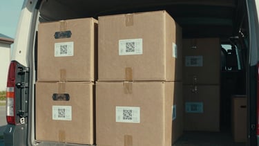 Cinematic documentary shot of humanitarian aid crates being unloaded from a vehicle in natural high-contrast daylight, with a visible QR code and tracking label on the side, cool-toned grading.