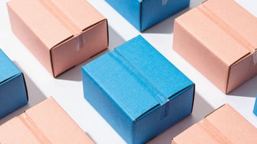 Overhead flat-lay of vibrant, candy-toned cardboard shipping boxes in electric peach and cobalt blue, hard direct sunlight with sharp playful shadows