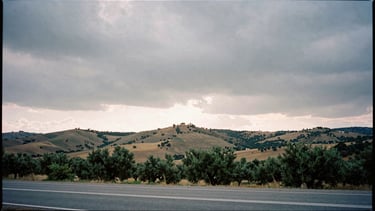 Wide documentary shot of the EX-370 road landscape at overcast sunset, rolling hills and olive groves under a heavy grey sky, high contrast 35mm film style