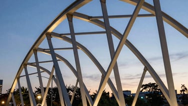 Modern urban park bridge with elegant steel arches at twilight in a North American city. The lighting highlights the geometric strength and aesthetic quality of the civil engineering work.