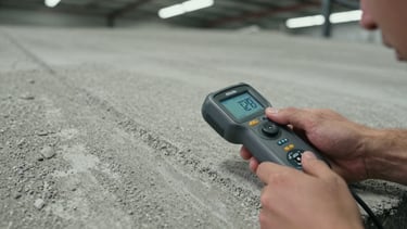 Close-up top-down shot of a technician's hands holding a concrete moisture meter pressed flat against a raw grey concrete floor surface, industrial warehouse ambient fluorescent overhead, texture of aggregate visible in the concrete, no text, no faces