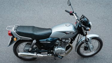 Overhead studio shot of a 150cc commuter motorcycle laid flat on a cool grey surface, instrument console and fuel tank badge in sharp focus, even neutral daylight from above revealing seat stitching and panel geometry