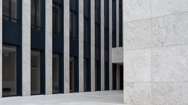 A minimalist architectural photograph of a high-end corporate building lobby in a North American metropolis. The space is defined by strong vertical lines of deep navy steel and expansive panels of light grey stone. The lighting is soft and natural, emphasizing a calm, professional atmosphere. Ultra-clean composition with significant whitespace.