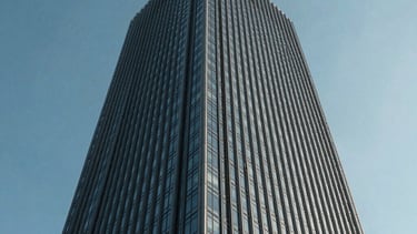A low-angle shot of a sleek corporate skyscraper against a clear, cool blue sky. The architecture is glass and steel, reflecting a sense of power and stability.
