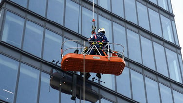 A heavy-duty rope suspended platform hanging securely against a modern glass skyscraper facade, crisp industrial photography, bright daylight, safety-orange accents, 35mm.
