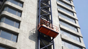 A high-capacity rack and pinion passenger hoist climbing the concrete side of a massive high-rise tower, bright daylight, navy blue steel frame, 35mm.