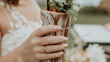A bride in a white wedding dress holds a tropical tiki cocktail mug garnished with fresh mint.
