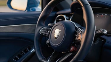 A close-up photograph of a high-performance European luxury car's cockpit, featuring hand-stitched leather in Deep Navy Blue and polished carbon fiber accents in Dark Charcoal. The steering wheel prominently displays a minimalist crest. Soft, natural evening light pours through the windshield, highlighting the precision of the stitching. North American / European Luxury Automotive Market setting.