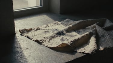 Cinematic, high-contrast 35mm film photograph of charcoal sketches and textured linen fabric folds on architectural polished concrete, dramatic low-key window light, deep shadows, desaturated warm tones