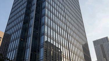 Clean, professional photography of a modern high-rise glass office building in a North American metropolitan business district during the afternoon. The lighting is crisp and natural, highlighting the architectural efficiency and sophisticated glass surfaces. The composition is a low-angle shot emphasizing growth and contemporary scale, utilizing a palette of deep navy and silver grey.